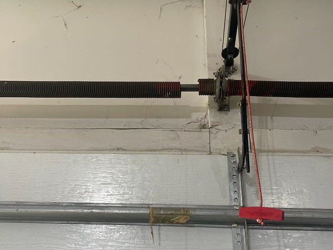 Torsion springs and shaft mounted across the header above a closed residential garage door, with the cable drums and the red emergency release cord in view